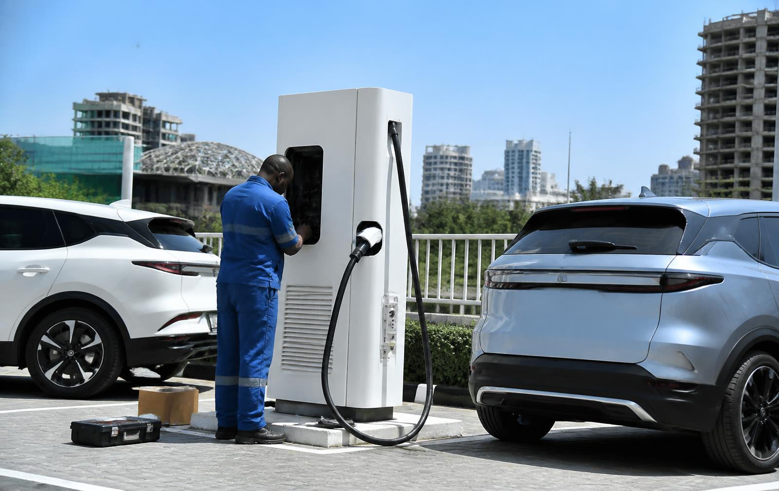 Technician installing a public electric vehicle fast-charging station in a Nigerian city car park with two EVs charging nearby